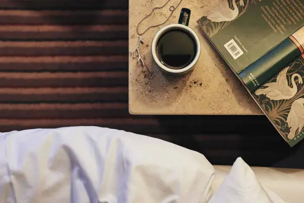 A coffee cup and an open book stand on a stone table. A white bedspread protrudes into the picture next to it.