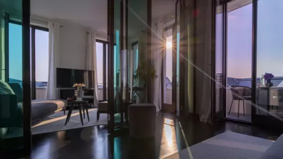 A modern hotel room on a sunny day. The sun shines through the open balcony door and casts a dazzling pattern of rays on the shiny wooden floor. The furnishings include a gray, curved couch and a matching stool, a small coffee table with flowers and fruit, and a flat-screen TV opposite the couch.  