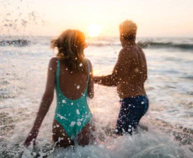 A woman in a turquoise swimsuit and a man in blue swimming trunks splash around in the sea at sunset. 