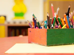 There is paper on a craft table and next to it is a colorful pencil box with crayons of different lengths. 