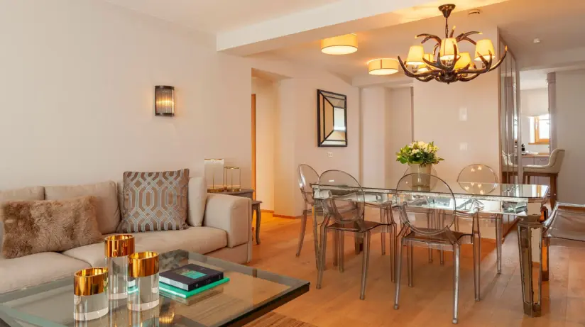 A brightly designed living and dining area with a beige sofa and a coffee table in the foreground. A dining table with 5 transparent chairs can be seen further back in the room.