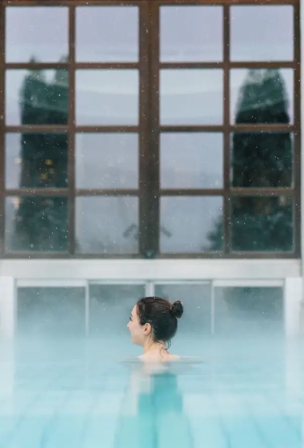 A woman swims in a warm, steaming indoor pool while fine snow falls from the sky. The entrance to the indoor pool can be seen in the background. 