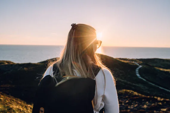 Eine Frau von hinten betrachtet, wie sie auf das Meer hinausblickt und den Sonnenuntergang genießt. Sie trägt eine Sonnenbrille, durch die der letzte Sonnenstrahl des Tages scheint, und hat einen Rucksack auf. Ihr Haar wird vom Abendlicht golden gefärbt. Der Weg vor ihr schlängelt sich durch die Küstenlandschaft.