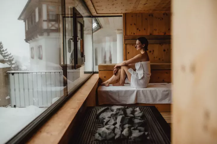 A woman is relaxing in a wooden sauna and overlooking the snowy landscapes through a big window front.