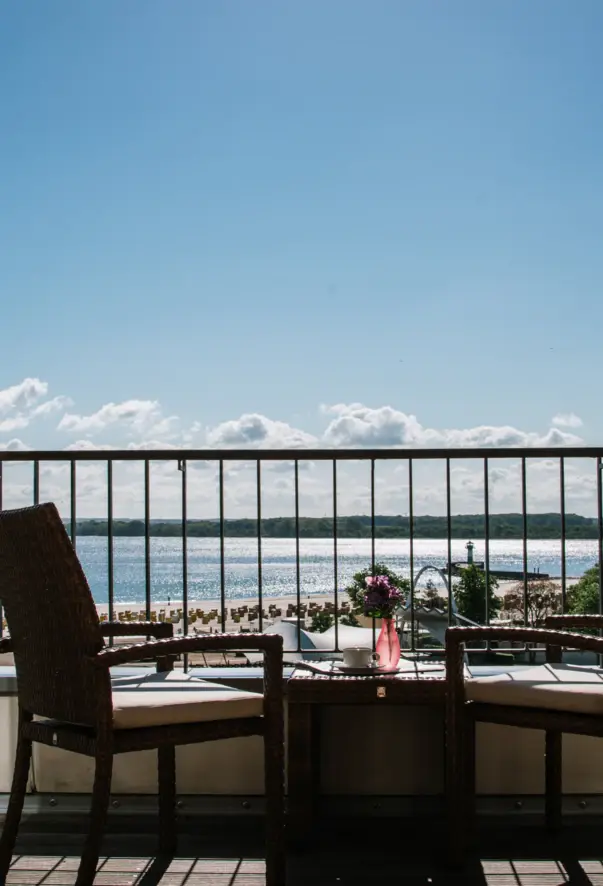 There are two chairs and a small table on a balcony overlooking the beach with beach chairs and the sea. 