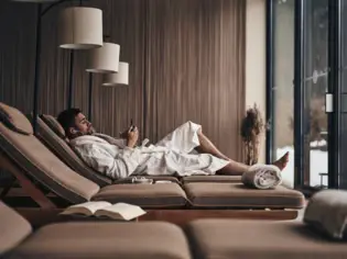 Man in a bathrobe lying relaxed on a lounge lounger in a relaxation area and looking at his smartphone.