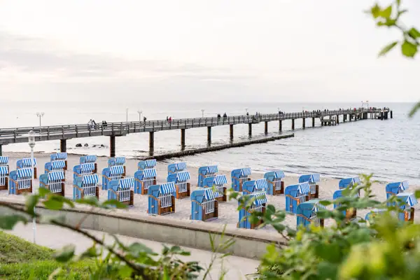 Strandkörbe am Strand mit einem Pier und Menschen im Hintergrund.