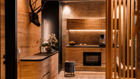 A wooden-look kitchen unit with a coffee machine and a fruit basket. A shelf with an orchid can be seen in the foreground and there is an artistic fake antler on the wall.