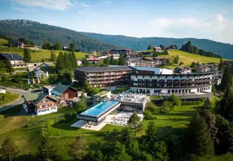 Building with a pool in the center of a mountain village, surrounded by trees and mountains.
