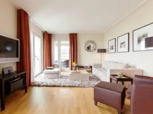 A spacious, bright living room with a beige sofa and orange curtains. Three pictures, a round mirror and a television hang on the walls. In front is a dark leather armchair with a footstool and in the background are large windows with a passageway to the loggia.