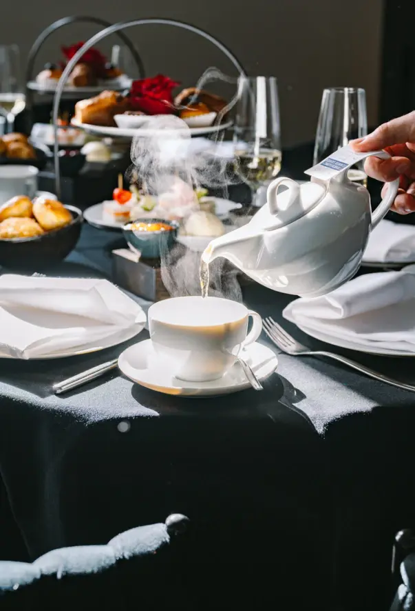 Tea is poured from a teapot into a white cup in front of a table laid with bread rolls and trays. 