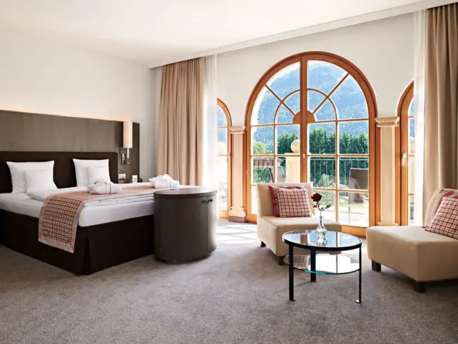 An elegant and cosy looking hotel room with a large bed and an Istzecke. The carpet is grey and daylight shines into the room through a large window