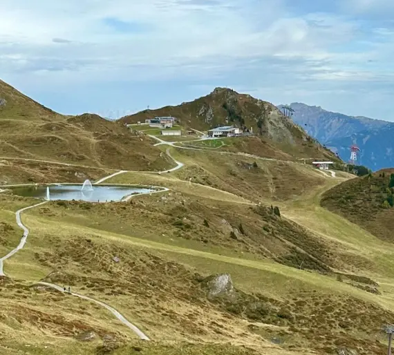 A path leads through a green landscape to a lake, surrounded by mountains and a cloudy sky.