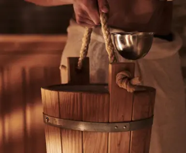 Person holding a wooden sauna bucket and a metal ladle in a wooden interior of a sauna.