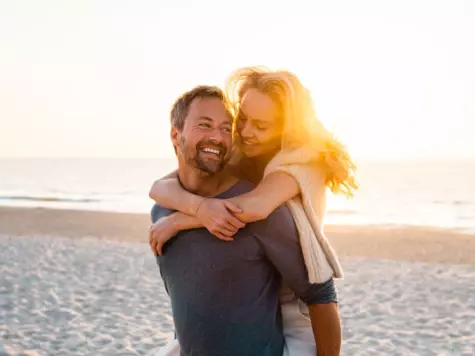 A man carrying a woman on his back on the beach whilst the sun is setting.