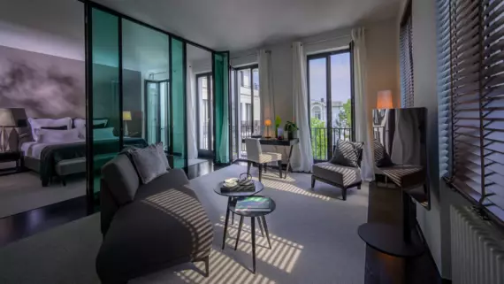 A luxuriously furnished hotel room divided into two areas by a glass wall. Part of the sleeping area can be seen on the left, with a large bed and dark-colored bed linen. On the right-hand side of the room, there is modern seating next to a table with a decorative vase and flowers. Natural light streams in through several floor-to-ceiling windows.