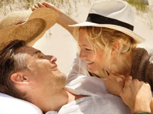 A couple wearing straw hats and lying happily together on the beach, smiling at each other. 