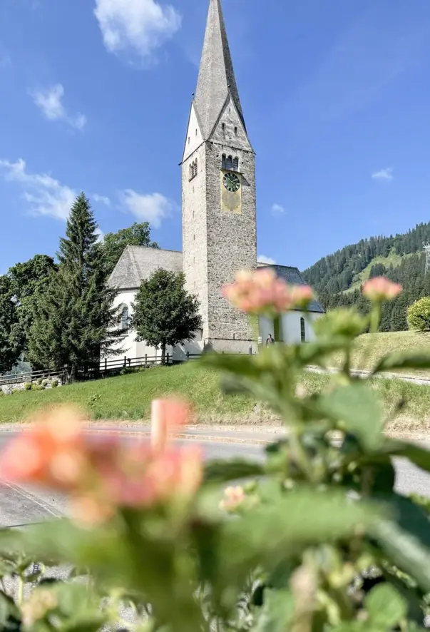 Historic church with striking bell tower and pointed roof against an Alpine backdrop in a summer landscape, with blurred flowers and green meadows in the foreground.