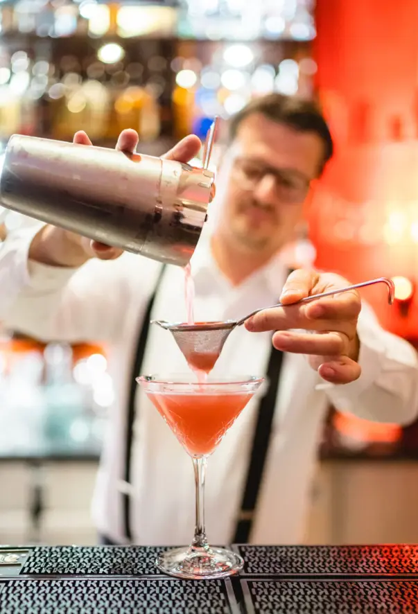 A bartender in professional attire concentrates on pouring a pink cocktail from a shaker through a strainer into a martini glass. 