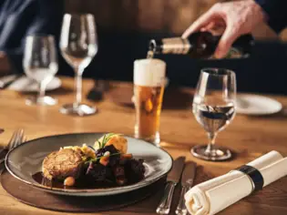 A plate with a main course stands on a laid wooden table and in the background a hand pours a beer into a beer glass.