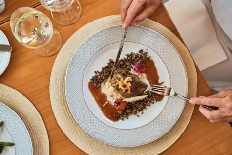 Person eats food on a plate, surrounded by cutlery and a wine glass.