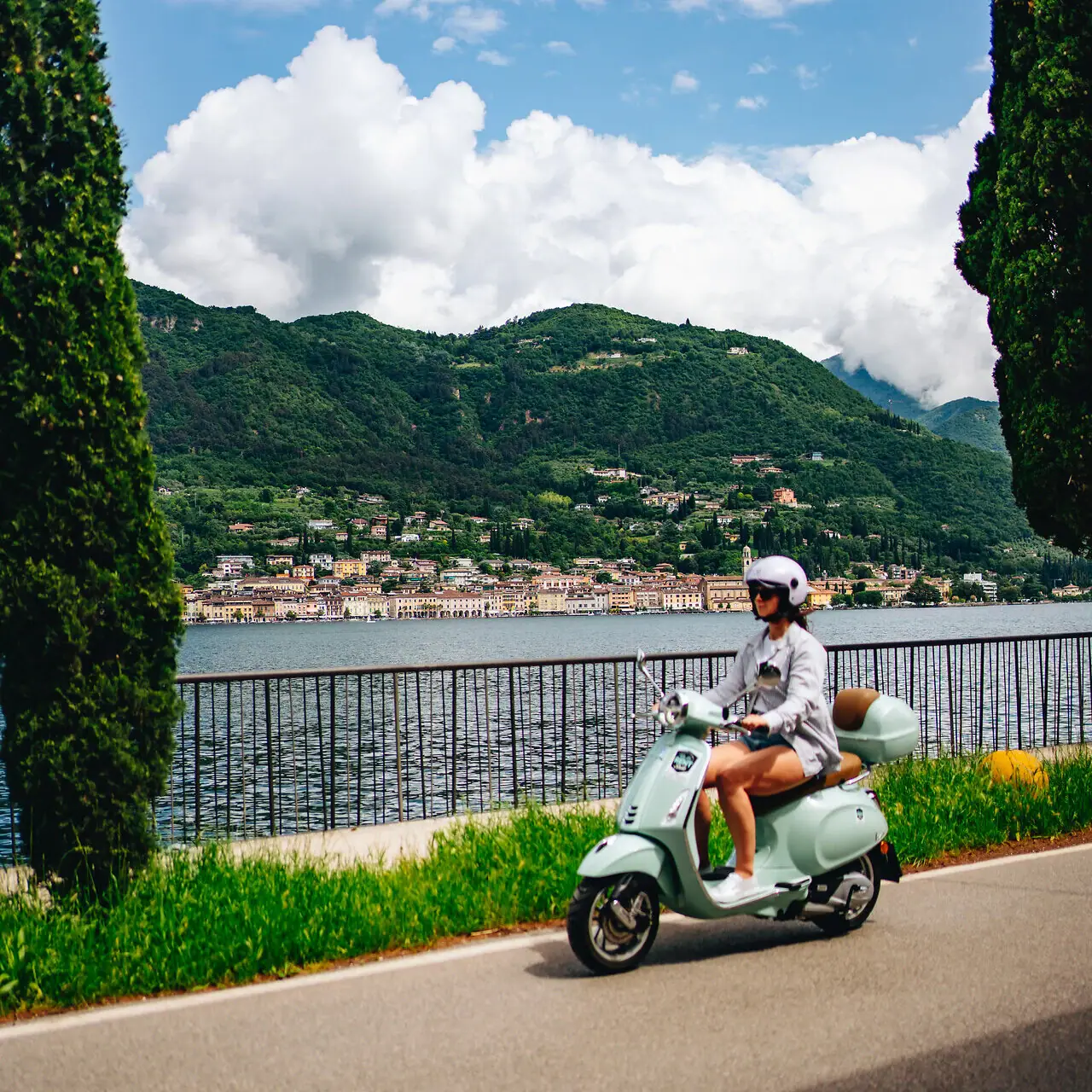 A woman riding a scooter on a road by a body of water.