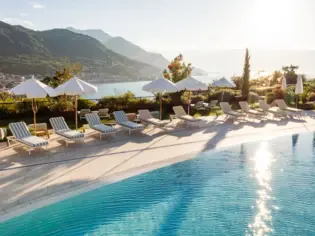 Pool with chairs and parasols by the water, surrounded by mountains and trees.