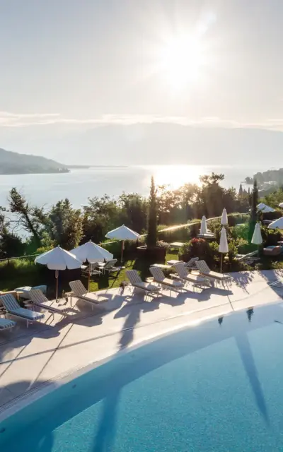 Pool with sun loungers and parasols on the banks of a body of water, surrounded by trees and mountains.