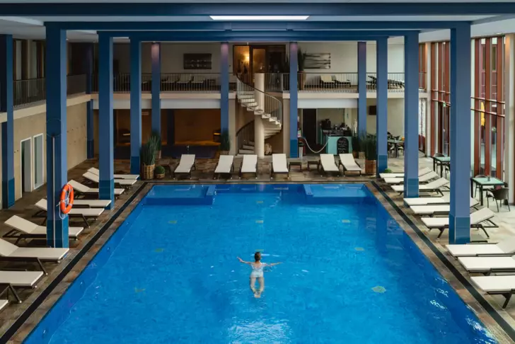 A person swimming in an indoor pool surrounded by white loungers. 