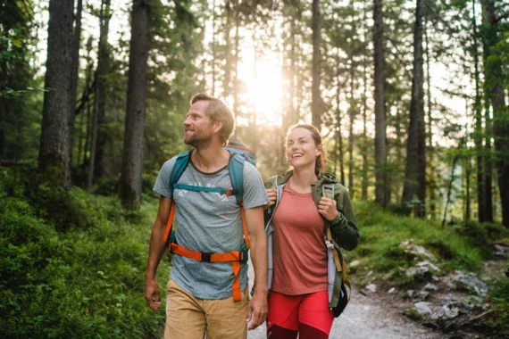 A couple in hiking gear is walking along a path in the forest while the sunlight is peaking through the trees. 