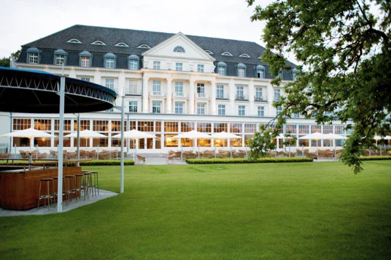 The photo shows the exterior of an elegant spa hotel with classic architecture. The multi-storey building has a white façade and a dark slate roof. In front of the building is a spacious terrace with plenty of seating and open parasols, providing a cozy atmosphere for al fresco dining. The terrace is surrounded by a well-tended lawn and the branch of a tree can be seen in the foreground, emphasizing the natural location. 