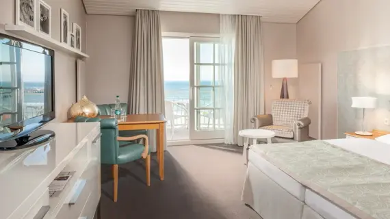 Inviting hotel room with a sea view, furnished with a double bed, the headboard of which is backed with wallpaper with a geometric pattern. In front of the floor-to-ceiling windows, which open onto a balcony with a view of the Baltic Sea, there are heavy gray curtains and a delicate drape. A cozy seating area with an upholstered armchair, a desk with green chairs and a large flat-screen TV complete the modern and comfortable furnishings. Black and white photographs are arranged on the wall above the desk, emphasizing the elegant atmosphere of the room.