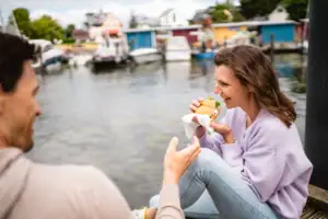 A woman sits on a jetty and eats a burger.