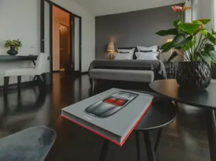 A modern bedroom with a large double bed in the middle. The bed is furnished with a gray comforter and several white pillows as well as a black decorative pillow. On either side of the bed are bedside tables, on each of which a table lamp with a silver base and white shade bathes the room in a warm light. The wall behind the bed is a dark shade of gray.