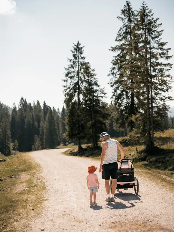A man and a child are walking along a gravel path, surrounded by trees.