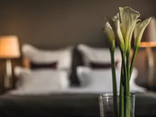 A vase of elegant white calla lilies stands in the foreground. A bedroom can be seen in the blurred background, illuminated by soft lighting from table lamps on either side of a double bed with white bed linen and black accent cushions.