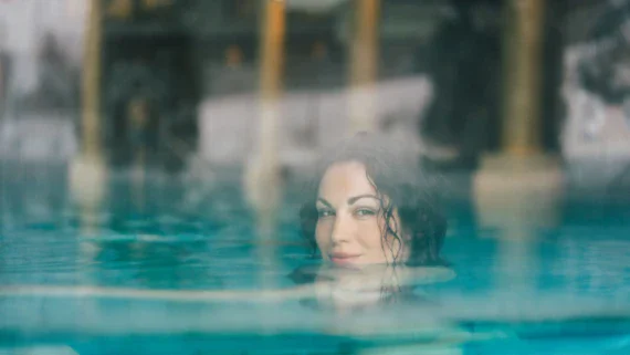 A woman is swimming chin deep in a warm, steaming pool and smiles. 