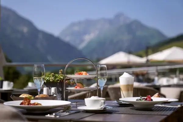 Tisch im Freien mit Geschirr, darunter Teller mit Essen und Getränken mit Bergen im Hintergrund.