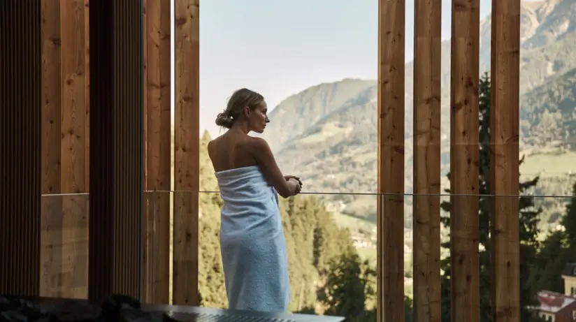 A woman stands on a balcony with a towel wrapped around her body and rests her hands on the glass balustrade.