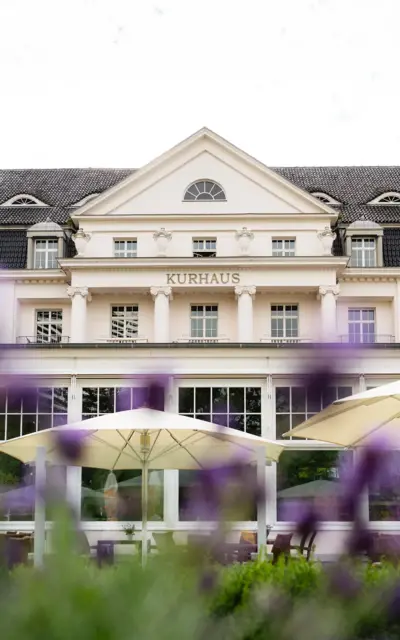 The white Kurhaus has an elegant style with columns on the second floor and wooden seating, shaded by white parasols. 