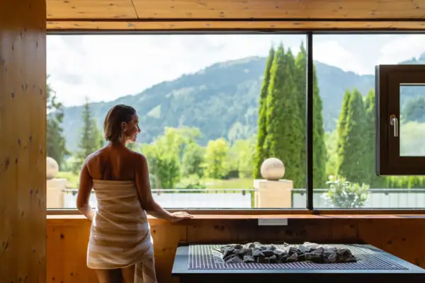 A woman standing indoors in front of a window, looking out to a mountain.