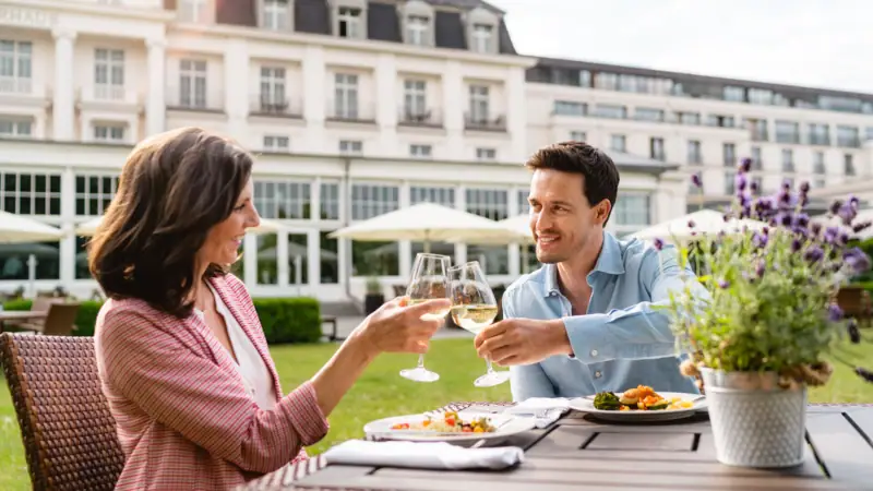 Ein Mann und eine Frau stoßen mit Weingläsern an einem Tisch im Freien an.