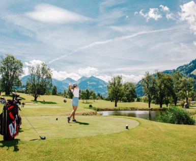 A woman stands on a golf course with her golf set and swings her club to take a shot. The course and the surrounding area are green and the sun is shining warmly.