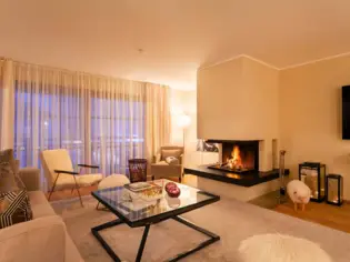 A bright living room with a fireplace and a sofa and two armchairs. In the middle is a large beige carpet with a glass coffee table on top.