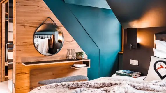 A sleeping area with a cozy bed in the foreground, on which lies a blanket and a decorative cushion. A stool can be seen behind the bed, standing in front of a small shelf and a round mirror.