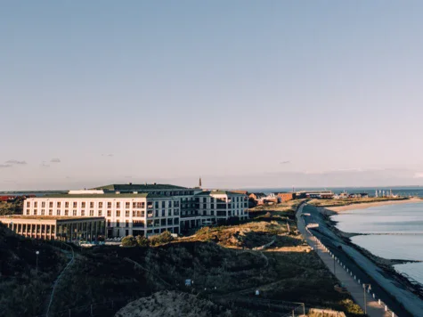 The white hotel complex of the A-ROSA Sylt surrounded by dunes and separated from the sea by a pedestrian path in the right part of the picture. 