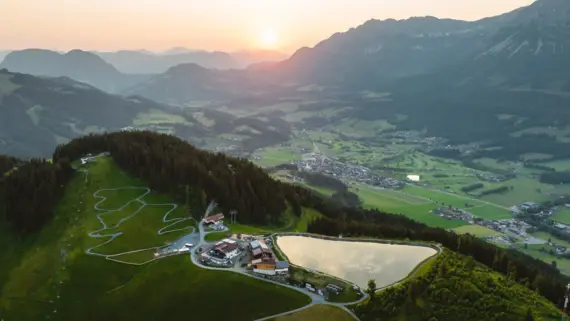 An aerial view of green mountains and a winding road along a lake. A valley with a small village can be seen in the background of the picture. 