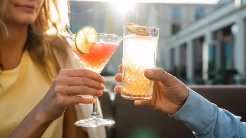 Zwei Personen stoßen bei Sonnenuntergang auf einer Terrasse mit Getränken an. Im Fokus steht eine Hand, die einen Cocktail mit Limettenscheibe in einem Martini-Glas hält, und eine andere Hand, die ein Highball-Glas mit einem klaren, sprudelnden Getränk und Orangenscheibe hält. Die warme Abendsonne beleuchtet die Szene, was eine entspannte und gesellige Stimmung vermittelt.