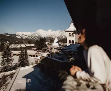 A woman stands on a balcony and looks out over a snow-covered landscape of houses, trees and mountains.