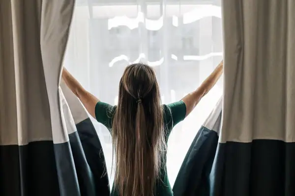 A woman opening curtains with her arms raised in a bright room.